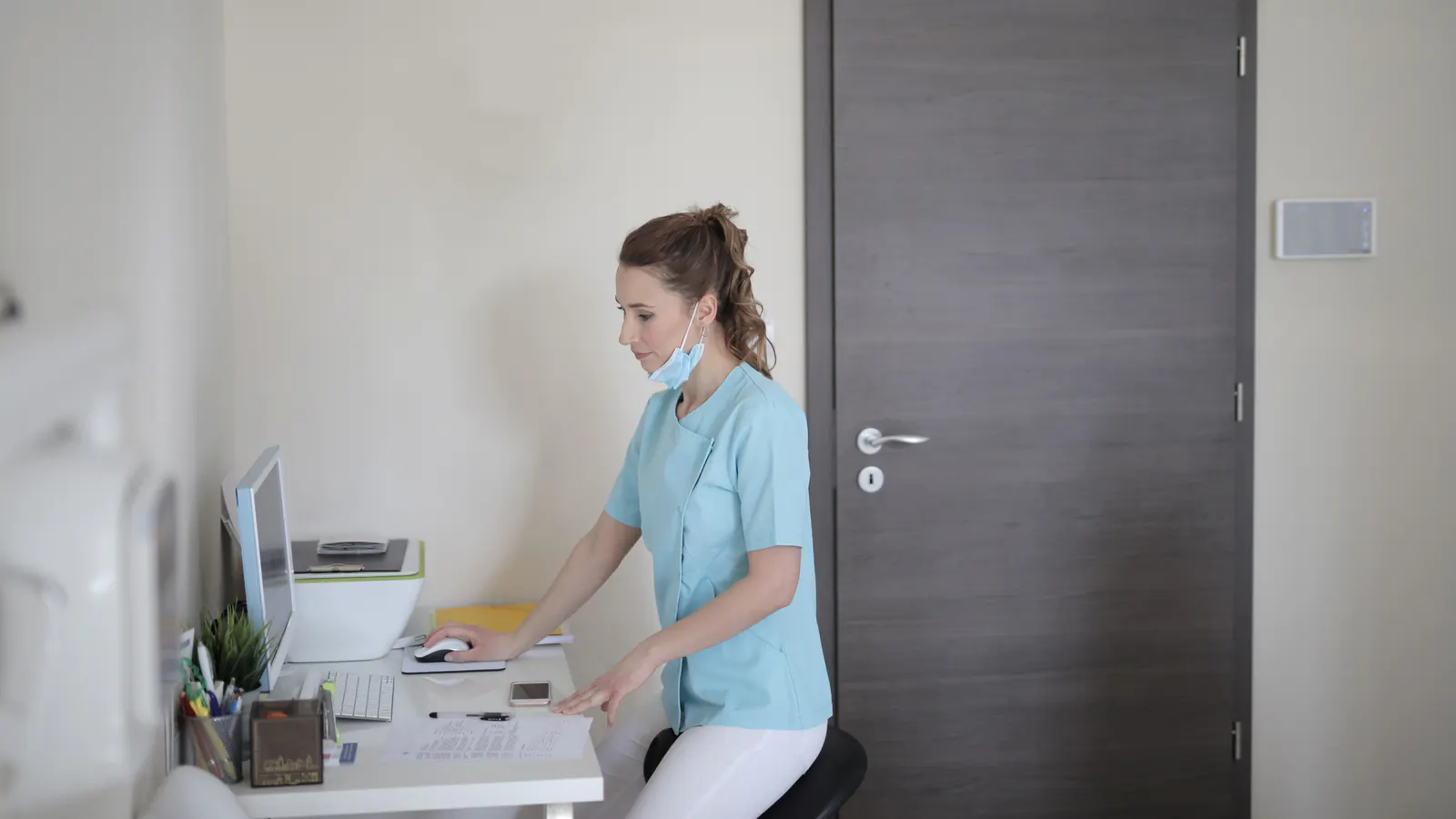 A clinic staff member in a light blue tunic works at a desk computer in a small office.