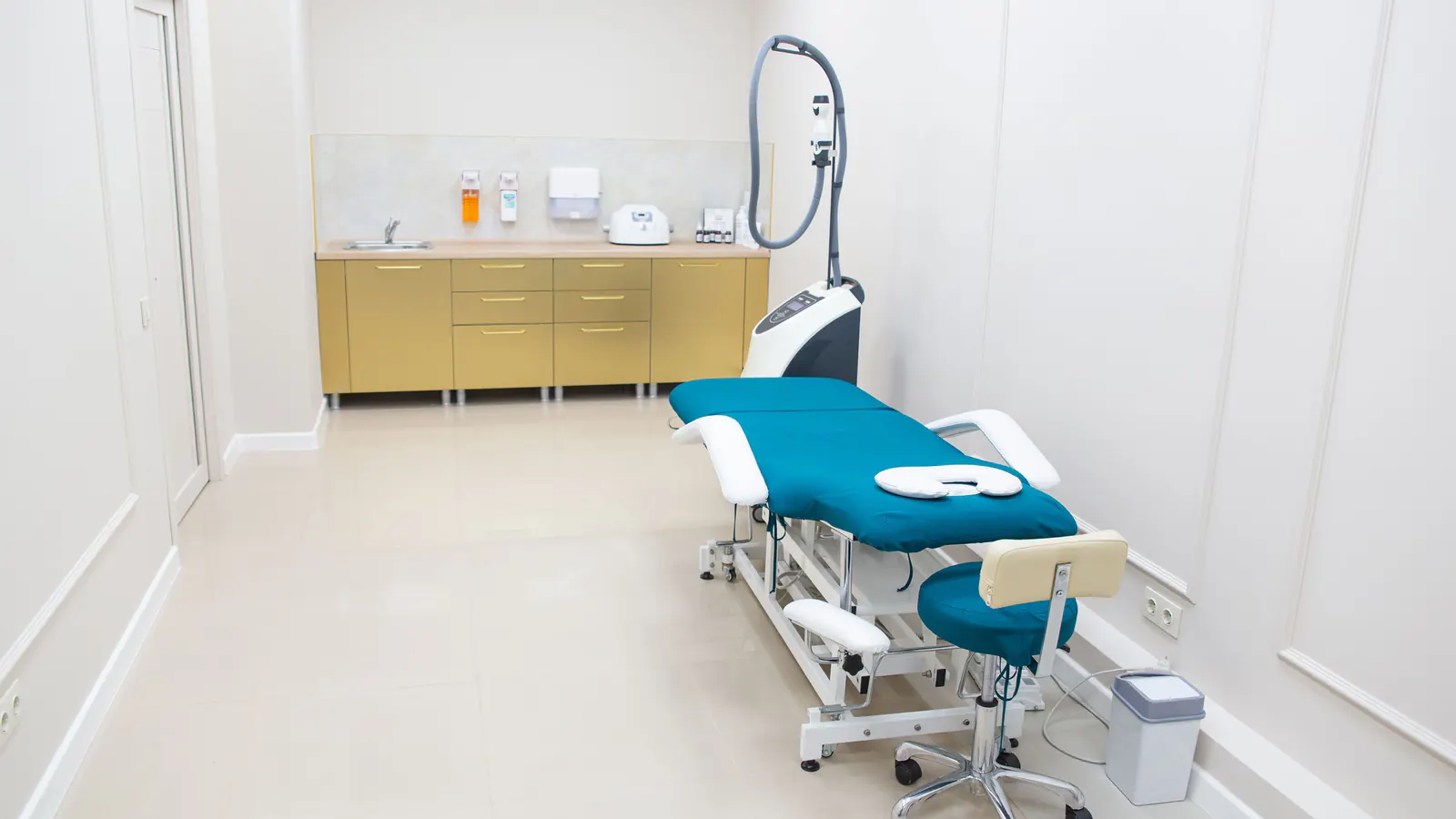 An empty med spa treatment room with a teal treatment bed, a stool and an aesthetic device on a stand.
