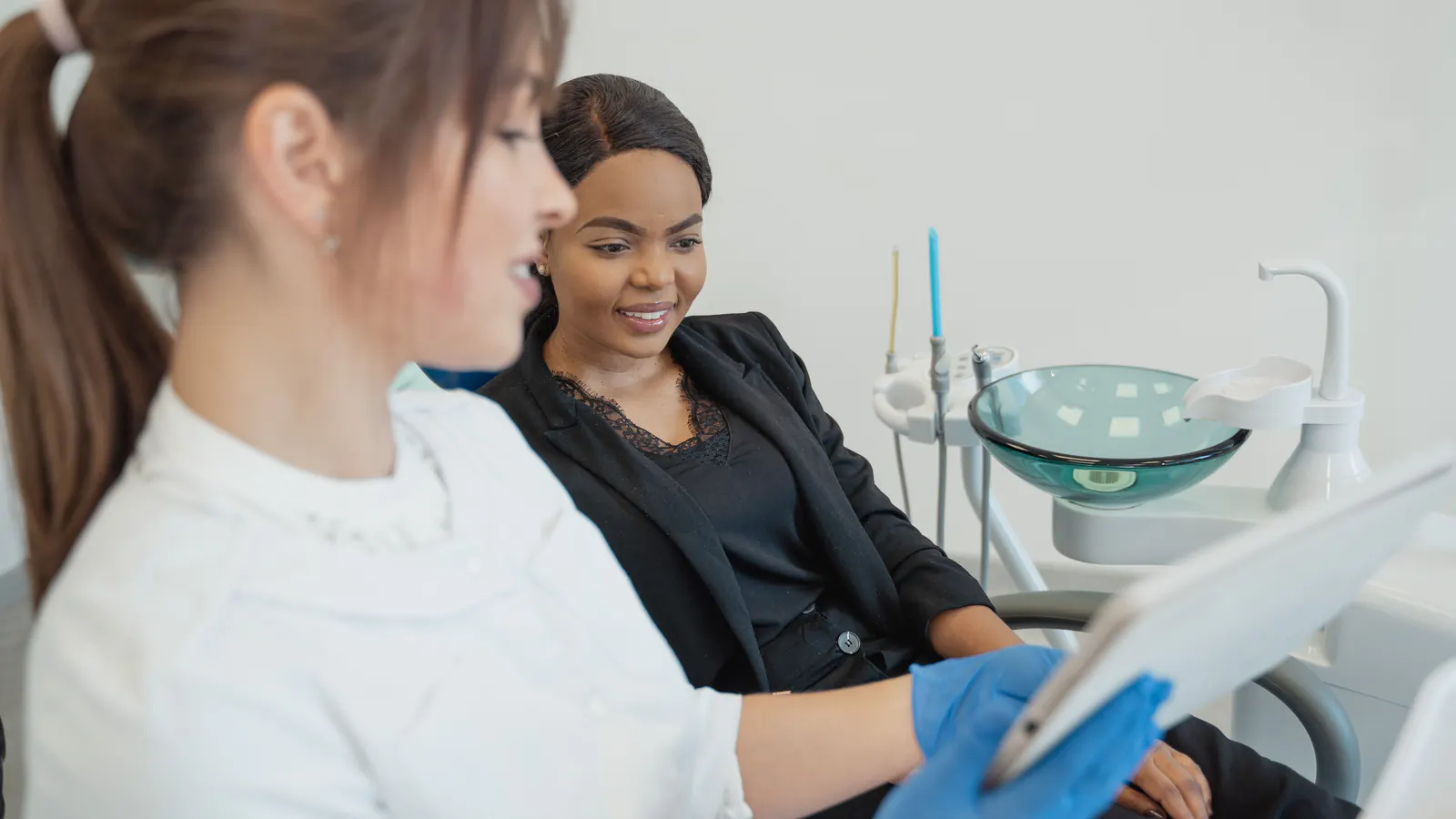 A treatment coordinator in blue gloves shows a smiling patient in a dental chair something on a tablet.