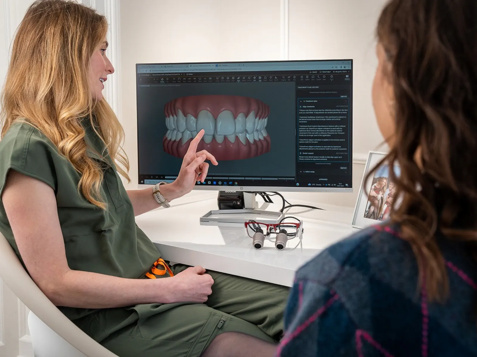A dentist points to a digital scan of a smile on a monitor while a patient watches during a consultation.