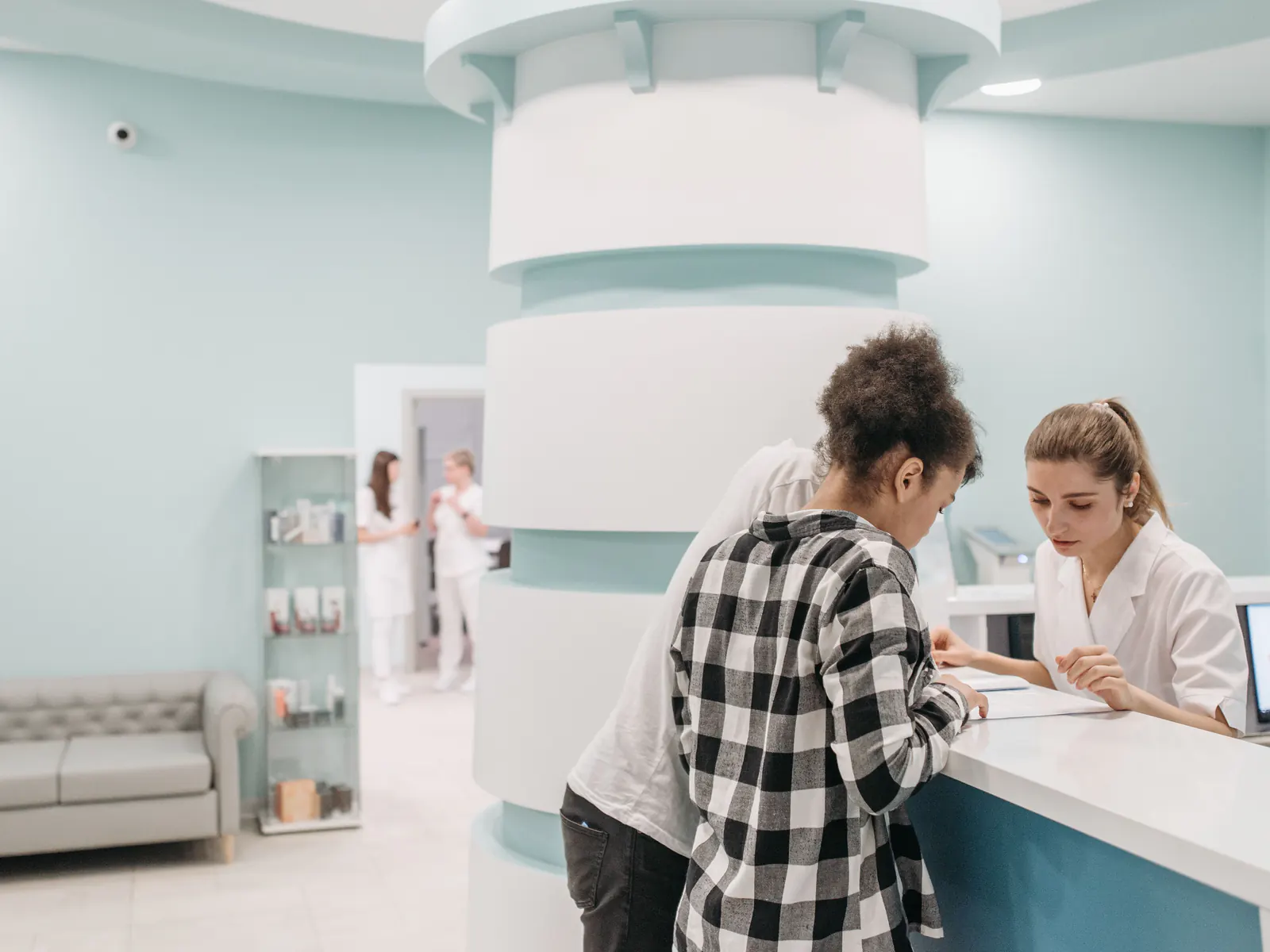 Two patients at a clinic reception desk while a receptionist in a white coat reads through a form with them.