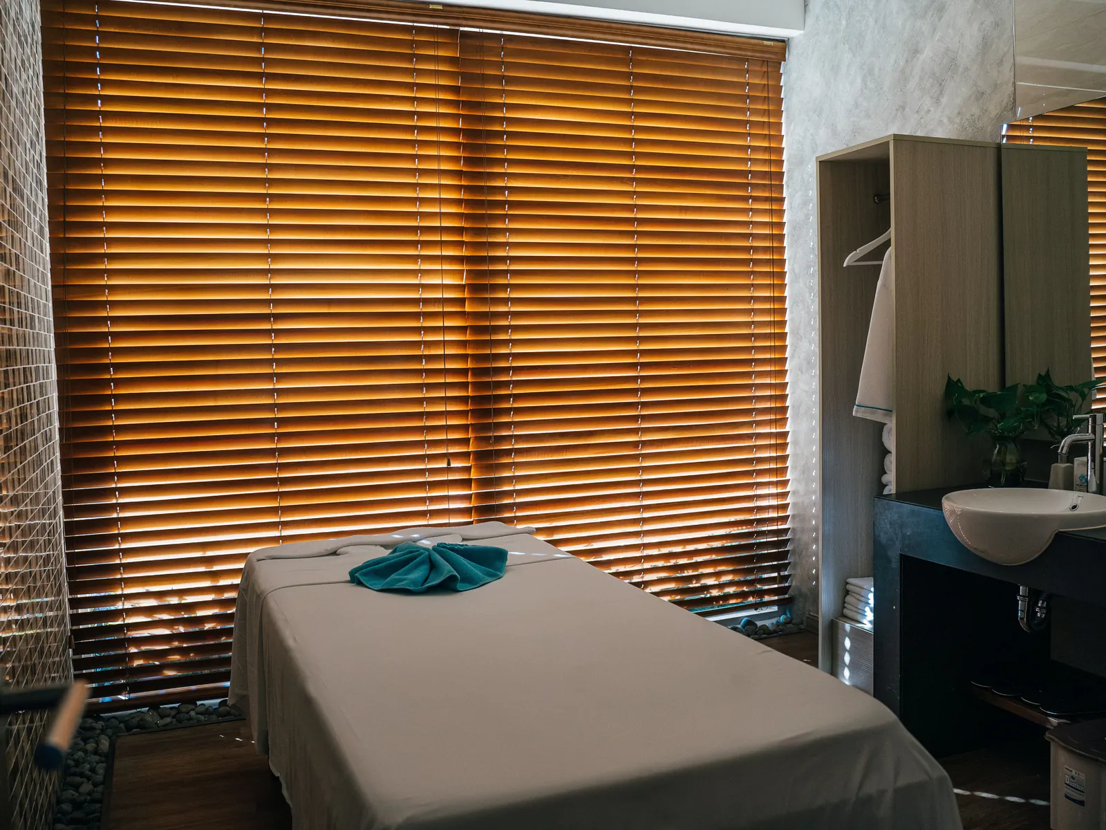 A massage room with a made-up treatment table and a folded towel, lit through wooden blinds.