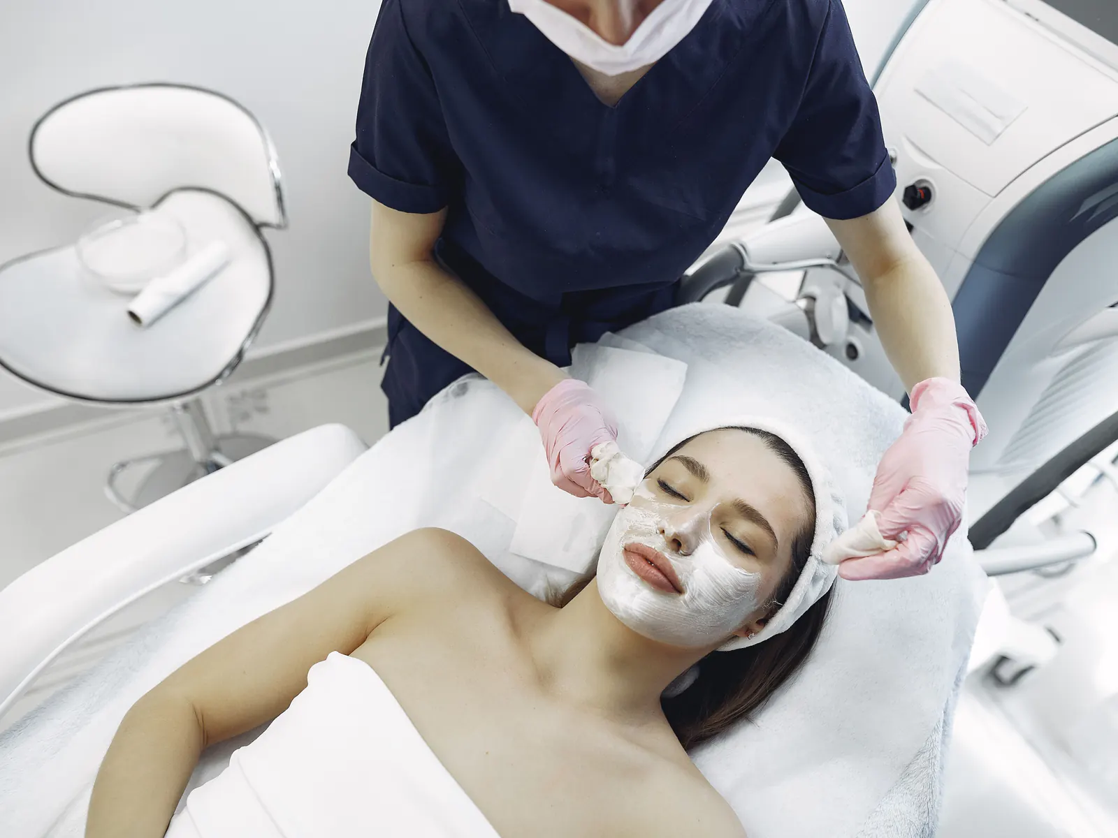 A practitioner in gloves applies a facial mask to a client lying on a treatment bed.