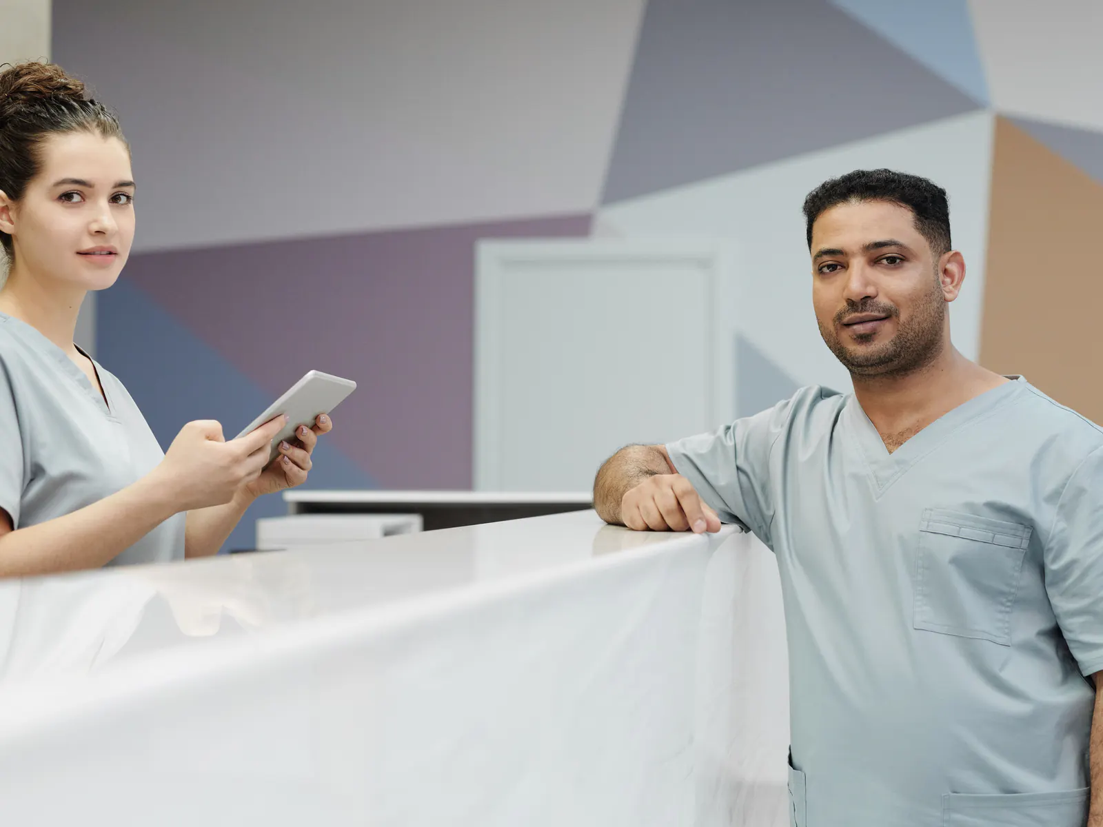 Two staff members in scrubs at a clinic front desk, one holding a tablet.