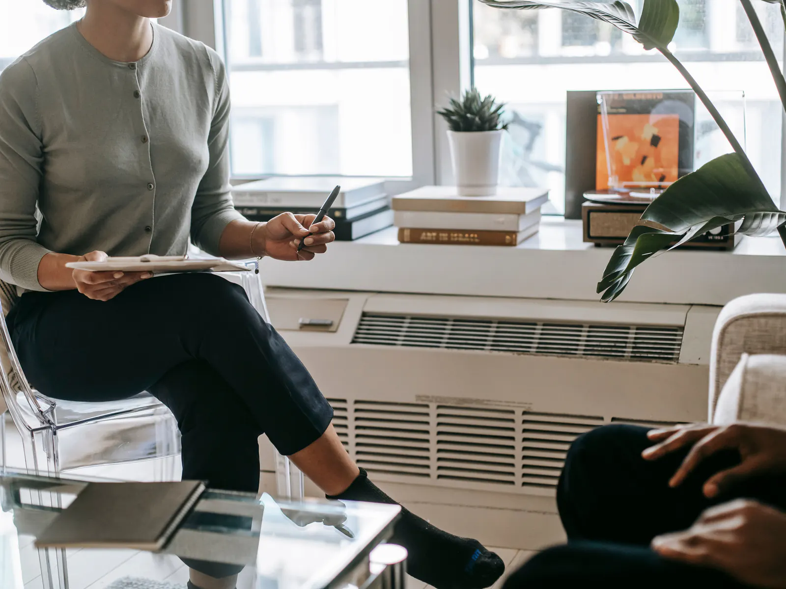 A therapist with a notepad and pen sits facing a client in a bright, quiet room, faces out of frame.