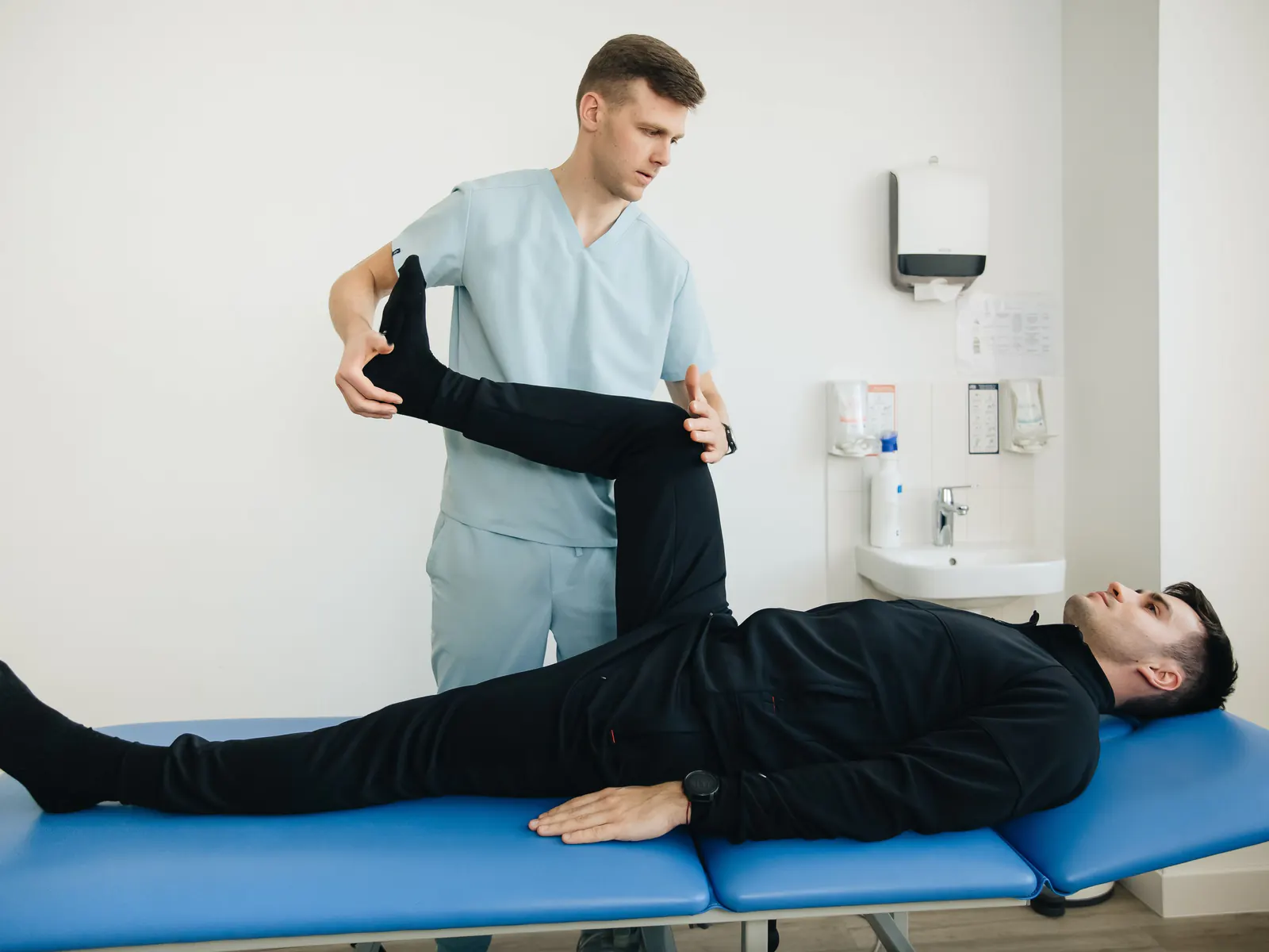 A physical therapist stretches the raised leg of a patient lying on a treatment table.